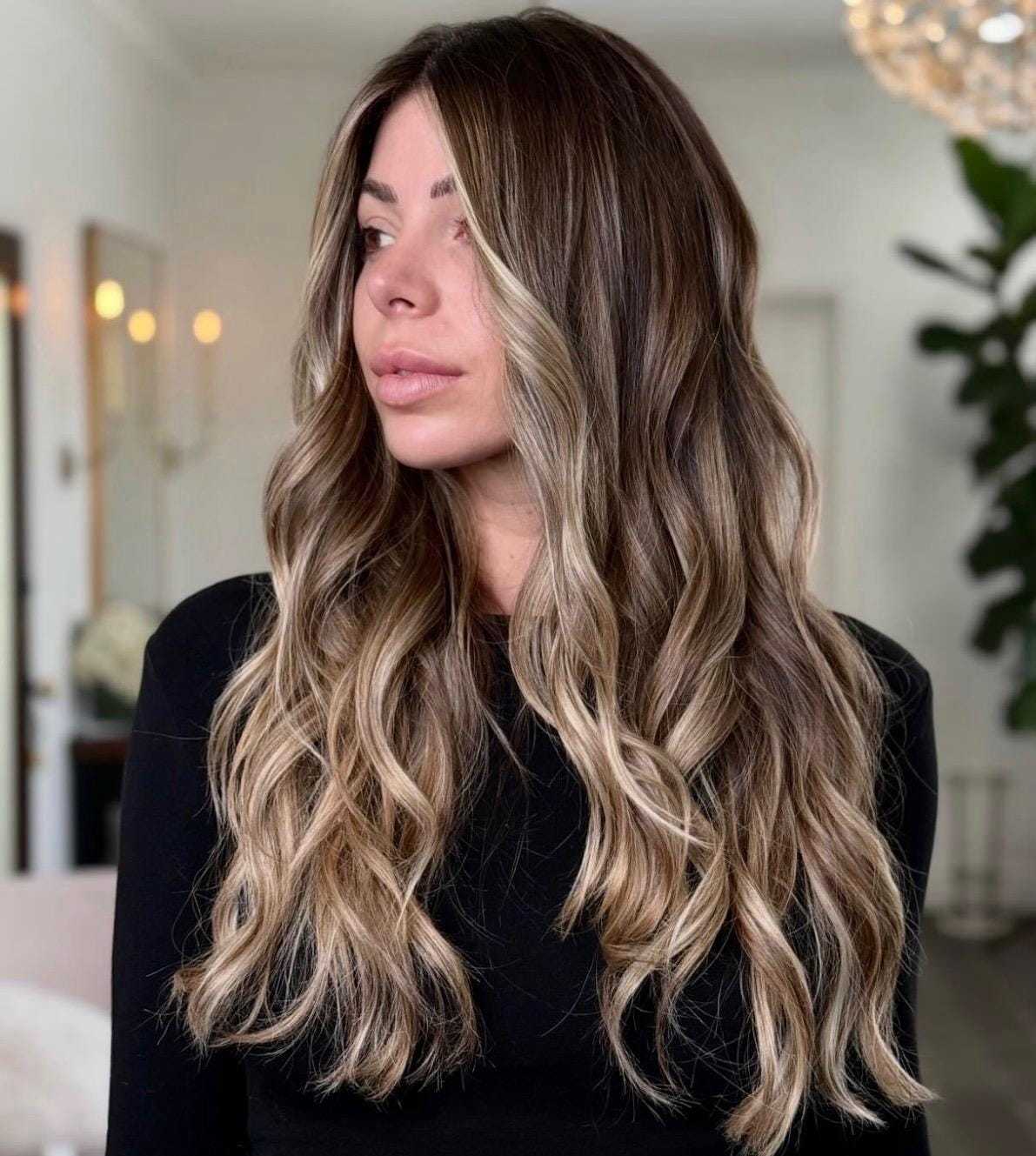 Long-haired woman with wavy, highlighted hair in stylish indoor setting.